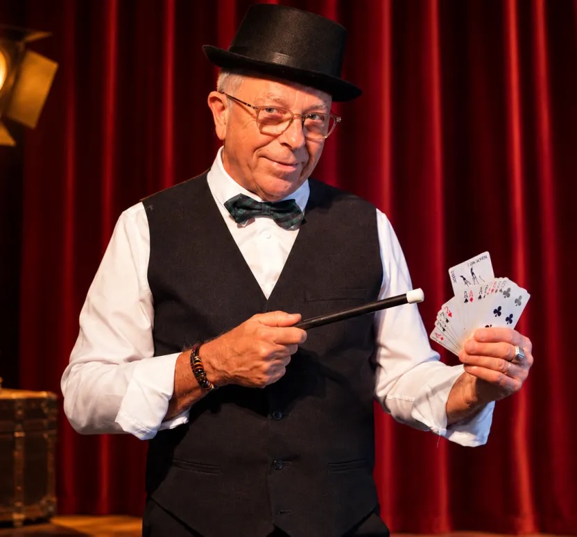 Philippe Archambault sur scène, baguette magique et jeu de cartes en main, devant un rideau rouge